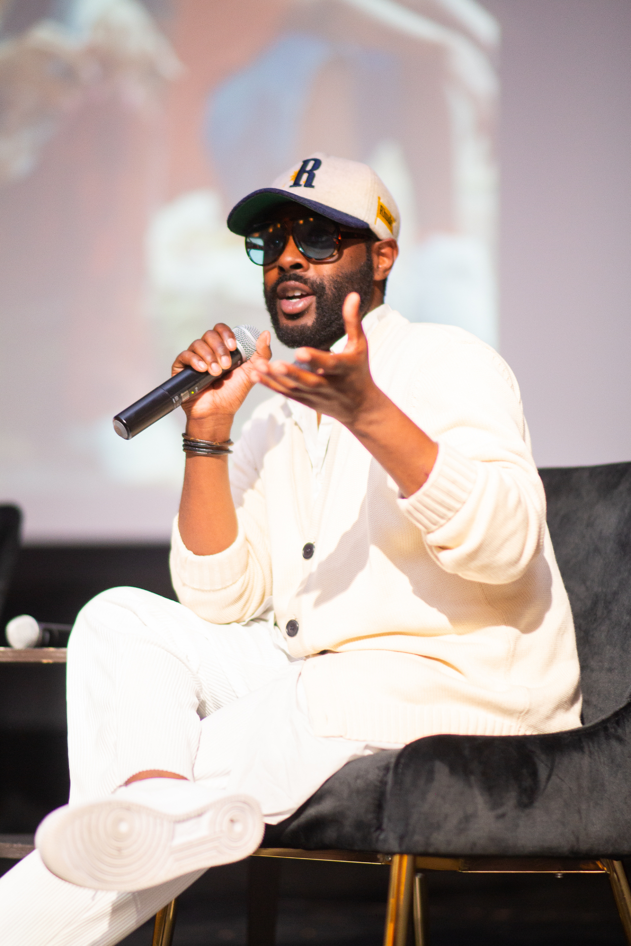 John Carmouche in a cap, tuxedo jacket and sunglasses, speaking into a microphone at a gathering